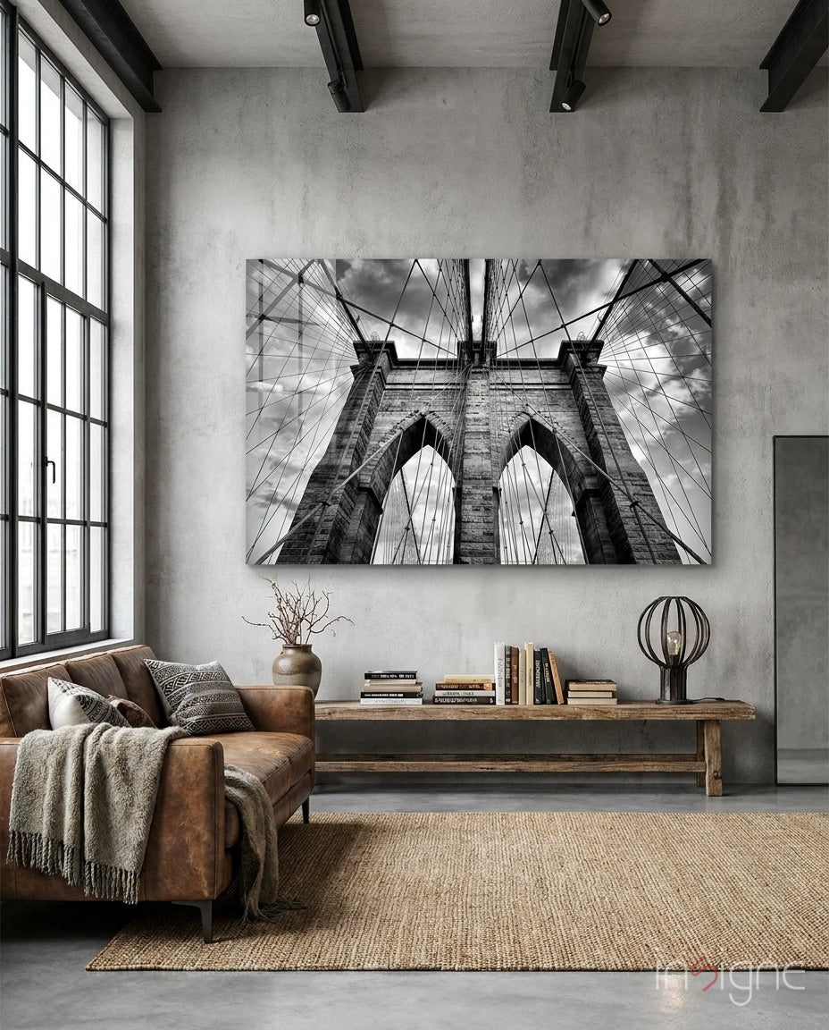 Brooklyn Bridge glass wall art displayed above a wood library bench in a concrete-walled industrial lounge with brown leather sofa and books