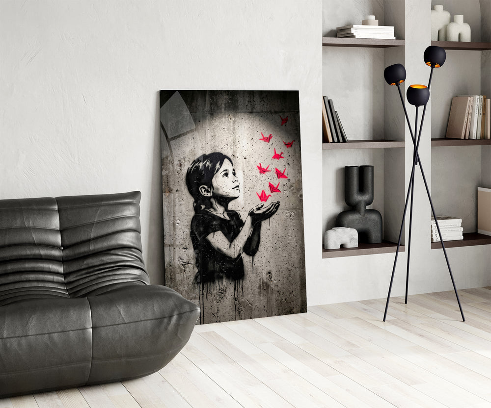 stencil girl releasing red paper cranes glass wall art leaning beside a dark leather sofa