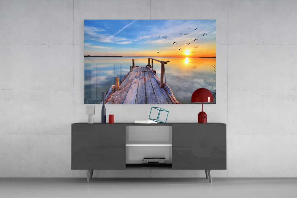 Fine art photography – worn wooden dock over still reflective lake at sunset, warm orange sky with birds taking flight on the horizon