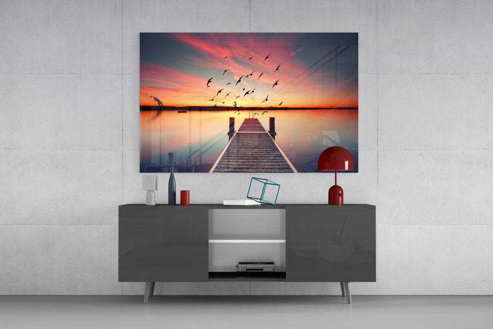 Fine art photography – wooden dock over mirror-still lake at dramatic orange sunset, large flock of birds sweeping across vivid sky