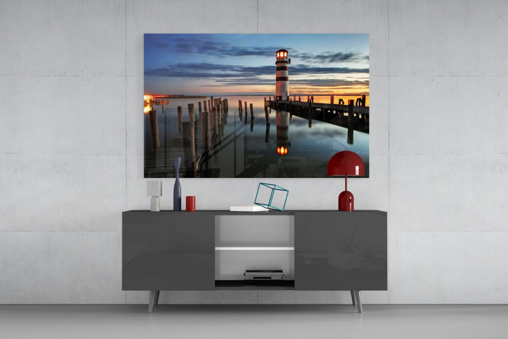 Fine art photography – striped lighthouse with glowing beacon at blue twilight, wooden pier in foreground, lamp reflected in calm lake