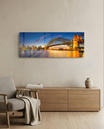 Panoramic harbor city at dusk, a great steel arch bridge lit gold beside a glittering downtown skyline mirrored in calm water. panoramic glass wall art A
