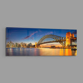 Panoramic harbor city at dusk, a great steel arch bridge lit gold beside a glittering downtown skyline mirrored in calm water. panoramic glass wall art E
