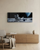 A spacesuited astronaut lounging in a deck chair on the lunar surface while planet Earth hangs in the black sky. panoramic glass wall art A