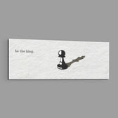 A single black chess pawn casting the long shadow of a king across a white textured surface, with a short motivational line of text panoramic glass wall art E