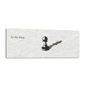 A single black chess pawn casting the long shadow of a king across a white textured surface, with a short motivational line of text panoramic glass wall art F