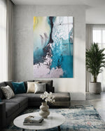 Colors of Abstraction glass wall art displayed above a charcoal sectional in a soft concrete-walled lounge with teal velvet pillow and palm vase