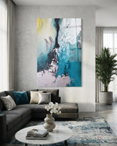 Colors of Abstraction glass wall art displayed above a charcoal sectional in a soft concrete-walled lounge with teal velvet pillow and palm vase