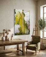 Colors of Abstraction glass wall art displayed beside a wood easel in a soft cream-plaster artist studio with green velvet armchair and olive tree
