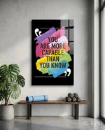 You Are More Capable glass wall art displayed above a wood bench in a soft white-plaster studio with monstera and yoga mat