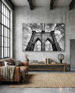 Brooklyn Bridge glass wall art displayed above a wood library bench in a concrete-walled industrial lounge with brown leather sofa and books