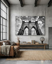 Brooklyn Bridge glass wall art displayed above a wood library bench in a concrete-walled industrial lounge with brown leather sofa and books