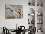 ginger kitten reaching a paw to a human hand glass wall art leaning beside a dark leather sofa