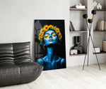 a blue-skinned woman crowned with sunflowers glass wall art leaning beside a dark leather sofa