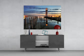 Fine art photography – striped lighthouse with glowing beacon at blue twilight, wooden pier in foreground, lamp reflected in calm lake