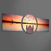 A wooden pier reaching into a calm lake at sunset with a flock of birds taking flight across a fiery orange sky. 3 piece glass wall art set F